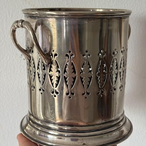 May include: A silver-colored wine bottle holder with a decorative openwork design. The holder has two curved handles and a circular base. The openwork pattern features leaf-like shapes. The holder is made of a shiny, reflective metal.