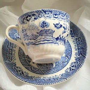 May include: A blue and white teacup and saucer set. The cup and saucer feature a detailed landscape design with pagodas, trees, and a boat. The set is made of porcelain and has a classic, vintage aesthetic.