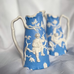 以下が含まれることがあります： 白のレリーフデザインが施された2つの青いセラミックピッチャー。ピッチャーは、白いハンドルとリムが付いた、質感のある青い表面が特徴です。1つのピッチャーには、木の枝に座っている人のレリーフがあります。もう1つのピッチャーも同様のデザインです。