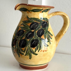 Può includere: Brocca in ceramica gialla con un motivo dipinto a mano di olive nere e foglie verdi. La brocca ha un bordo e una base marroni ed è adatta per servire bevande.