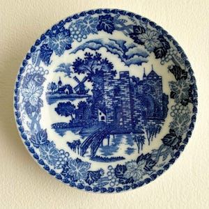 Może przedstawiać: Okrągły, biało-niebieski talerz dekoracyjny. Talerz przedstawia szczegółową niebieską scenę krajobrazową z zamkiem, mostem i wiatrakiem. Obrzeże zdobi niebieski wzór winogron i liści.