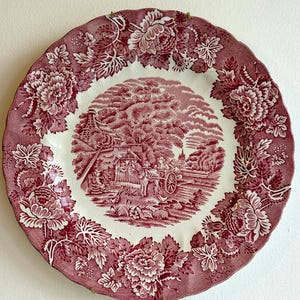 May include: A round, antique-style plate with a red and white floral design. The plate features a central scene of a rural landscape with a horse-drawn cart, surrounded by a border of large floral patterns. The plate has a scalloped edge.