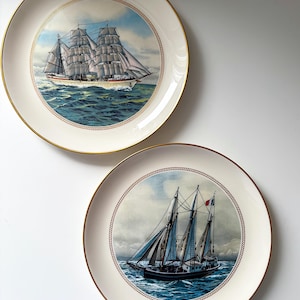 Può includere: Due piatti decorativi a tema nautico. I piatti hanno uno sfondo color crema e bordi dorati. Uno presenta una grande nave a vela con vele bianche su un oceano blu. L'altro mostra una barca a vela più piccola.