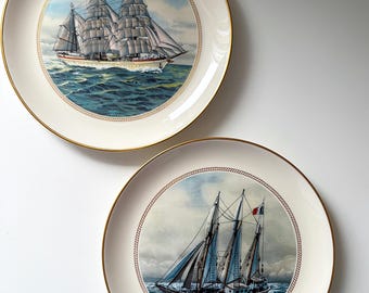 May include: Two decorative plates with nautical themes. The plates have a cream-colored background with a gold rim. One plate features a large sailing ship with billowing white sails on a blue ocean. The other plate shows a smaller sailboat.