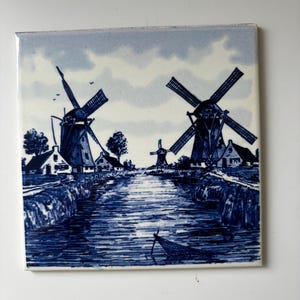 May include: A square ceramic tile featuring a blue and white illustration of a canal scene with two large windmills, smaller houses, and a small boat in the foreground. The artwork uses varying shades of blue to depict the water, sky, and structures.
