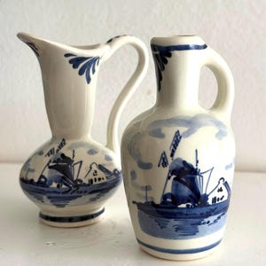 May include: Two white ceramic vases with blue windmill and landscape designs. One vase has a handle and a curved spout, the other has a rounded body and a flared top. Both are decorated with blue floral accents.