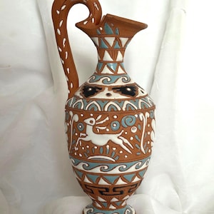 May include: A terracotta-colored ceramic vase with a curved handle and a wide spout. The vase is adorned with white, blue, and black geometric and animal designs. The handle has white dot accents.