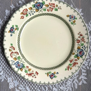 Könnte beinhalten: Cremefarbener Keramikteller mit gewelltem Rand. Er hat ein Blumendesign mit blauen, roten und gelben Blumen, grünen Blättern und einem grünen Rand. Der Teller hat einen Durchmesser von ca. 25 cm.