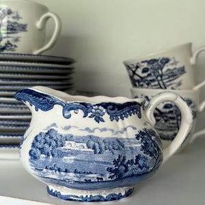Könnte beinhalten: Eine weiß-blaue Keramik-Sahnekännchen mit Landschaftsdesign. Das Sahnekännchen hat einen gebogenen Griff und einen Ausguss. Weitere passende Tischartikel, darunter Tassen und Teller, sind im Hintergrund gestapelt.