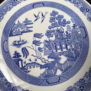 May include: A blue and white ceramic plate with a Willow pattern design. The plate features a detailed landscape scene with pagodas, bridges, and birds. The rim has a decorative border. A classic piece for serving or display.