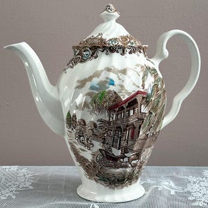 May include: White ceramic teapot with a detailed brown and green transferware scene of a building and horse-drawn carriage. The teapot has a curved spout, a loop handle, and a decorative lid with a finial.