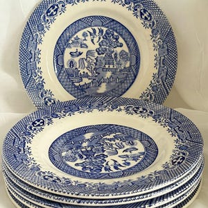 Puede incluir: Un conjunto de platos de cerámica azules y blancos con un diseño de patrón Willow. Los platos presentan una escena central de paisaje rodeada por un borde decorativo. Los platos están apilados.