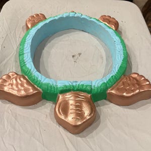 Puede incluir: Una maceta con forma de tortuga con acabado color cobre en las patas y la cabeza. El cuerpo de la maceta es un anillo con interior azul claro y exterior verde. La cabeza de la tortuga tiene un diseño detallado.