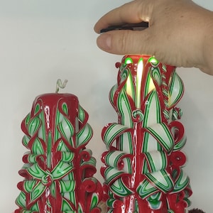 Könnte beinhalten: Zwei geschnitzte rote Kerzen mit grün-weißen bandartigen Designs. Die Kerzen sind angezündet, mit einer Hand, die ein Feuerzeug über einer hält. Die Kerzen stehen auf einer reflektierenden Oberfläche, mit getrockneten Blumen im Vordergrund. Festlich und dekorativ.