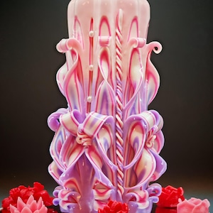Peut inclure: Une bougie sculptée ornée de rose, violet et blanc, avec des motifs floraux et en forme de ruban. De petites bougies rouges en forme de fleur entourent la base. La bougie a un aspect décoratif et artistique.
