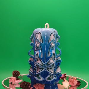 Könnte beinhalten: Eine geschnitzte Kerze in Blau-, Weiß- und Türkistönen mit aufwendigen blütenartigen Designs. Die Kerze steht auf einer verspiegelten Oberfläche mit getrockneten Blumen und Grün, vor einem einfarbigen grünen Hintergrund. Die Kerze hat einen Docht oben.