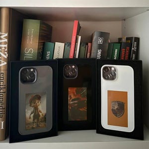 Puede incluir: Tres fundas para smartphone exhibidas en cajas negras sobre una estantería con libros. Las fundas tienen diferentes diseños: un personaje de dibujos animados, una escena de coche y un emblema de Porsche sobre fondo naranja. Las fundas son plateadas, negras y blancas.
