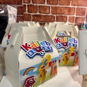 Puede incluir: Cajas blancas con forma de cabaña con asas, decoradas con personajes de dibujos animados coloridos, arcoíris y el texto "Kaileb Blocks". Las cajas tienen un esquema de color azul, rojo y blanco, perfectas para una fiesta.