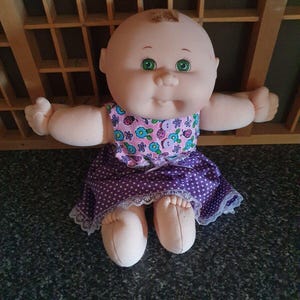 May include: A vintage doll with a soft, rounded face and green eyes. The doll wears a sleeveless dress with a floral top and a purple polka-dot skirt trimmed with lace. The doll's arms are outstretched.