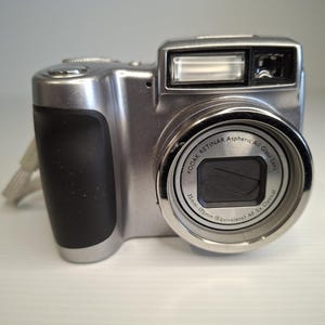 May include: A silver digital camera with a black rubberised grip on the left side. The lens barrel features text reading Kodak Retinar Aspheric All Glass Lens 35mm-175mm (Equivalent) AF 5x Optical. A rectangular flash unit is positioned above the lens.