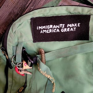 May include: A green backpack with a black patch that reads "IMMIGRANTS MAKE AMERICA GREAT" in white lettering. The backpack has black zippers, straps, and a key ring with keys and a red tag.