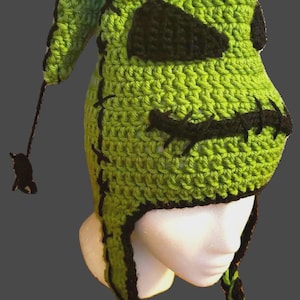 May include: A green and black crocheted hat with the text "PATTERN ONLY!". The hat features a face design with black stitched details and long braided tassels. The hat is displayed on a white mannequin head.