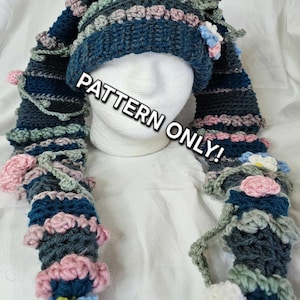 Könnte beinhalten: Handgefertigte Häkelmütze mit langen Ohrenklappen, verziert mit rosa, blauen und weißen Blumenakzenten. Die Mütze ist hauptsächlich dunkelblau mit grauen und grünen Streifen. Der Text "PATTERN ONLY!" ist sichtbar.