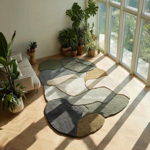 Puede incluir: Una alfombra multicolor con un diseño orgánico en forma de piedras. La alfombra presenta tonos de verde, gris, beige y marrón. Está colocada sobre un suelo de madera en una habitación con grandes ventanales y varias plantas en macetas.
