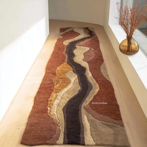 Puede incluir: Una alfombra larga y rectangular con un diseño similar a un río. La alfombra presenta una franja central marrón oscuro, flanqueada por capas de secciones beige, arena y óxido. La alfombra está colocada sobre un suelo de madera clara.