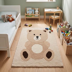 Op de afbeelding: Een rechthoekig vloerkleed met een groot, centraal beige teddybeerontwerp met een crèmekleurige buik, donkerbruine voetzolen en een gezicht met eenvoudige zwarte stiksels. Het kleed heeft een zachte, pluizige textuur op een lichte houten vloer.