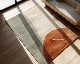 Könnte beinhalten: Ein rechteckiger Teppich mit modernem Design. Der Teppich hat ein Farbblockmuster mit einem großen halbkreisförmigen Bereich in Rostorange, einem hellbeigen Abschnitt und einem gedämpften grünen Abschnitt. Der Teppich liegt auf einem Holzboden.