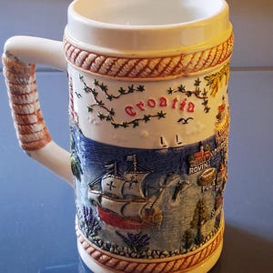 May include: White ceramic beer stein with a detailed relief design. The stein features a nautical scene with a ship, the word "Croatia", and various coastal town illustrations. The handle is rope-textured.