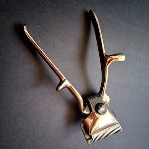 May include: A vintage manual hair clipper constructed from polished silver-toned metal. The tool features two long, curved handles that operate a spring-loaded mechanism, connected to a rectangular cutting head with fine, serrated metal teeth.