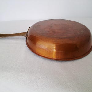 May include: A copper frying pan with a long, brass handle. The pan is round with a curved rim and a shiny, reflective surface. The handle is attached to the side of the pan.