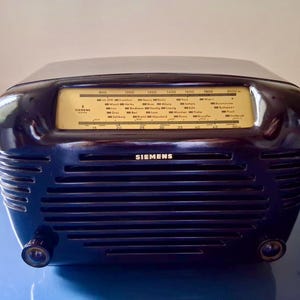 May include: A vintage black Siemens radio with a curved top and a horizontal speaker grill. The radio dial is yellow with black text and a tuning needle. The word "SIEMENS" is printed on the front.
