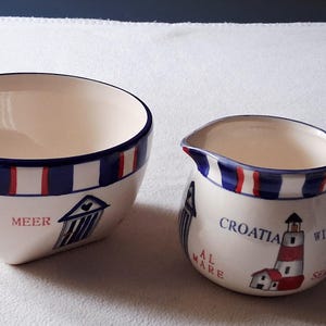Op de afbeelding: Een keramische kom en creamer set met een nautisch thema. De kom en creamer zijn wit met blauwe en rode strepen rond de randen. De creamer heeft een vuurtoren illustratie en het woord "Croatia".