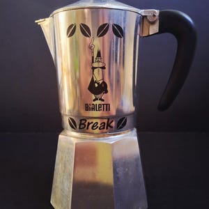May include: A silver-coloured aluminium stovetop espresso maker with a black handle and lid knob. The body features a black graphic of the Bialetti mascot with one arm raised, the text "Bialetti Break," and stylised leaf icons printed on the front.
