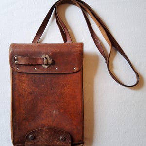Puede incluir: Bandolera de cuero marrón vintage con una larga correa para el hombro. La bolsa rectangular presenta un cierre de solapa con un broche metálico y remaches. El cuero muestra signos de desgaste, lo que aumenta su estética antigua. La bolsa está diseñada para llevarse al hombro.