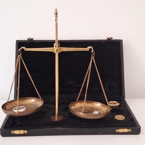 May include: A brass balance scale with two hanging pans suspended by chains from a central beam. The scale is positioned in an open black case with a velvet-lined interior. A silver ring sits in the left pan, and small metal weights are in the right.
