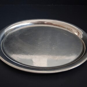 Peut inclure: Un plateau de service ovale de couleur argentée polie avec une surface lisse et réfléchissante et un bord légèrement surélevé. Le plat en métal présente un design simple et épuré avec une large bordure et un centre plat, brillant.