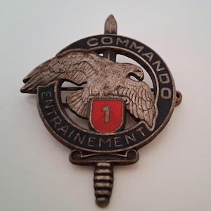 May include: A silver-toned metal military badge featuring a central eagle perched above a red shield with the number 1. The emblem is surrounded by a circular black border with the text COMMANDO ENTRAINEMENT and a vertical dagger design.