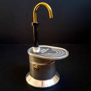 Puede incluir: Una cafetera de aluminio plateado para estufa con un surtidor vertical negro rematado por una boquilla de metal dorado curvado. La base es cilíndrica con una pequeña válvula color latón, y la plataforma superior tiene grabado el texto mini express.