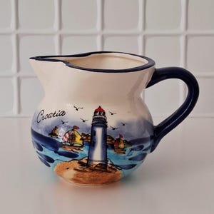 May include: A cream-coloured ceramic pitcher featuring a hand-painted coastal scene with a lighthouse, sailboats, and the word Croatia. The pitcher has a dark blue handle and a matching dark blue rim, with a rounded body and a wide pouring spout.