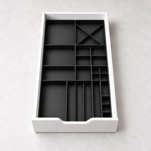 Puede incluir: Un organizador de cajones rectangular blanco con un interior negro dividido en múltiples compartimentos de varios tamaños, incluyendo secciones cuadradas, rectangulares y en forma de cruz para guardar objetos.