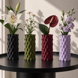 May include: Four cylindrical vases with a geometric diamond-patterned exterior, coloured black, olive green, deep red, and light purple. Each vase holds a different floral arrangement, including a white lily, daisies, a red anthurium, and purple flowers.