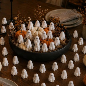 May include: A collection of small white ghost figurines with simple black eyes and mouths, arranged in a large black bowl filled with orange and white pumpkins and dried foliage, with additional ghost figurines scattered across the table.