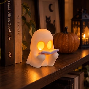 May include: A white, glowing ghost-shaped lamp sits on a wooden shelf, holding a small open book. The lamp features two illuminated oval eyes and emits a warm light. It is positioned next to a textured orange pumpkin and a black metal lantern.