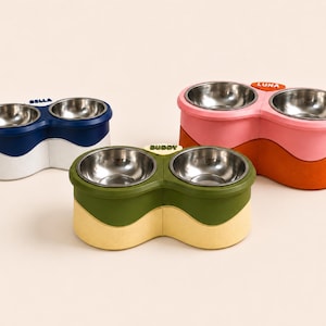 Pode incluir: Três estações duplas de tigelas para animais com inserções de aço inoxidável. Uma é azul-marinho e branca com o nome BELLA, uma é verde-oliva e amarela com o nome BUDDY, e uma é rosa e laranja com o nome LUNA. Cada uma tem um design ondulado.