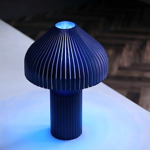 May include: A dark blue table lamp with a mushroom-shaped shade and a cylindrical base, both featuring vertical slats. The lamp emits a bright blue light from the top opening and the base, casting a blue glow onto the surface below the lamp.