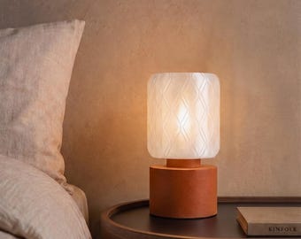 May include: A table lamp featuring a cylindrical terracotta-colored base and a translucent white glass shade with a subtle embossed diamond pattern. The lamp emits a warm glow, illuminating the textured surface of the glass shade.
